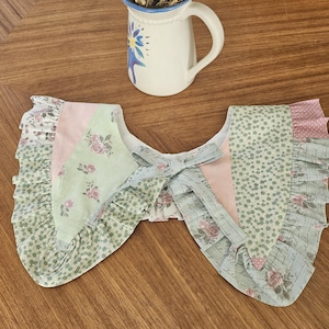 Könnte beinhalten: Ein pastellfarbener Patchwork-Kragen mit Schleife. Der Kragen hat ein Blumenmuster in Rosa, Grün und Weiß. Der Kragen ist wahrscheinlich handgefertigt und verleiht einen Vintage-Look.