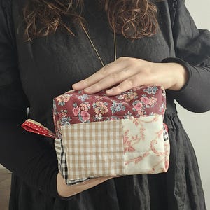 Könnte beinhalten: Patchwork-Stoffbox-Tasche mit floralem Muster oben, einem beige-weiß karierten Muster in der Mitte und einem rot-weiß gepunkteten Griff. Die Tasche wird von einer Person in einem schwarzen Kleid gehalten.