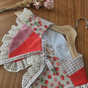 Pode incluir: Um babete feito à mão com estampa de morango, xadrez vermelho e acabamento em renda. O babete está em um cabide de madeira. O babete tem uma gravata borboleta e é feito de tecido. O fundo é uma superfície de madeira.
