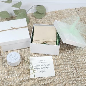 May include: A white gift box with a beige drawstring pouch inside, a separate white lid with a light green tulle bow, a small white circular speaker, and a square card that reads: EASTER May your heart be light and your day be bright.