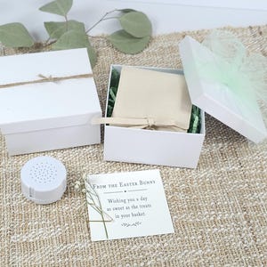 May include: Two white square gift boxes, one closed with twine and one open containing a beige fabric pouch with a ribbon. A small white circular speaker, a square card with the text From the Easter Bunny Wishing you a day as sweet as the treats in your basket, and a sprig of dried greenery are included.