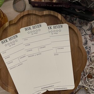 May include: Two off-white rectangular book review cards featuring the printed text BOOK REVIEW, five outline stars for rating, and fields for title, author, started, finished, and thoughts and quotes, with the handle @oldladyaesthetics at the bottom.