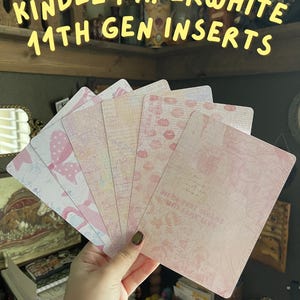 May include: A hand holds a fan of decorative Kindle Paperwhite 11th Gen inserts. The inserts feature pink and white patterns, including bows, lips, and floral designs. The text "KINDLE PAPERWHITE 11TH GEN INSERTS" is displayed at the top.