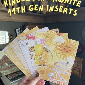 May include: A hand holding a fan of decorative Kindle Paperwhite 11th Gen inserts. The inserts feature yellow and white designs with illustrations of insects, flowers, and inspirational quotes. The text "KINDLE PAPERWHITE 11TH GEN INSERTS" is at the top.