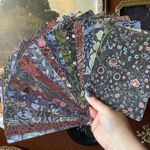 May include: A fan of rectangular cards featuring diverse botanical and floral patterns in shades of dark green, muted pink, blue, and brown. Each card displays intricate, repeating nature-inspired illustrations with dense foliage and flower motifs.