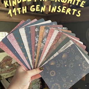 May include: A hand holds a fan of Kindle Paperwhite 11th Gen inserts. The inserts feature various patterns, including floral, celestial, and abstract designs in shades of pink, blue, green, and brown. Text at the top reads "KINDLE PAPERWHITE 11TH GEN INSERTS".