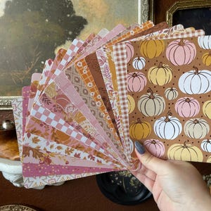 May include: A fan of rectangular paper cards featuring various autumn-themed patterns including pumpkins in shades of white, pink, and yellow on a brown background, alongside checkered, floral, and leaf prints in warm orange, pink, and brown tones.
