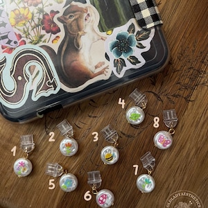 May include: A collection of eight small, round, white pearl-like charms, each featuring a unique colorful graphic such as a flower, bee, or butterfly. Each charm is attached to a clear plastic clip by a small gold-toned metal jump ring.