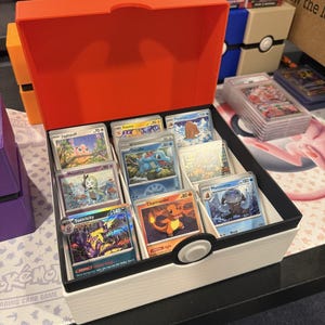 May include: A white and orange plastic storage box with a black trim and a circular button design on the front, filled with various trading cards including Toxtricity, Charmander, Mamoswine, Totodile, Jigglypuff, Meloetta, Piloswine, and Pzema.