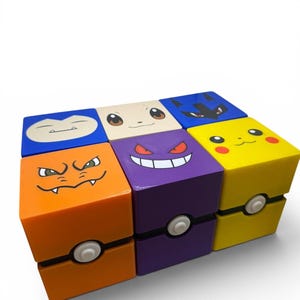 May include: A set of six square storage boxes arranged in two rows. Each box features a different character face on top and a Poké Ball design on the front, including orange, purple, yellow, blue, and cream colours with various expressions.