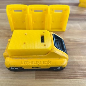 May include: A yellow DeWalt 20V Lithium Ion 3Ah battery pack positioned in front of three yellow plastic battery wall mounts. The battery features black accents, a rectangular shape, and clearly visible text indicating its voltage and capacity.