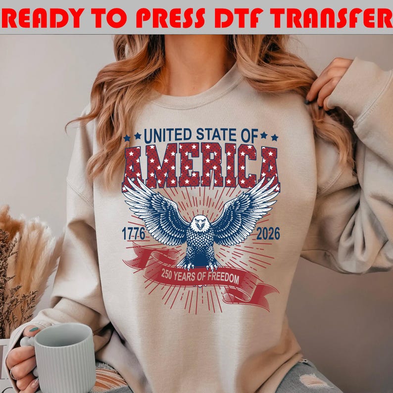 Op de afbeelding: Een beige sweatshirt met een grafische print met de tekst UNITED STATE OF AMERICA in het rood met witte sterren. Daaronder staat een blauwe adelaar met gespreide vleugels, de jaartallen 1776 en 2026 en een rood lint met 250 YEARS OF FREEDOM.