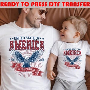 Op de afbeelding: Twee witte t-shirts met een patriottische afbeelding van een Amerikaanse zeearend, met rode en blauwe tekst: UNITED STATE OF AMERICA, 1776, 2026 en 250 YEARS OF FREEDOM. Het ontwerp bevat een rood lint en sterpatronen in de dikke letters.