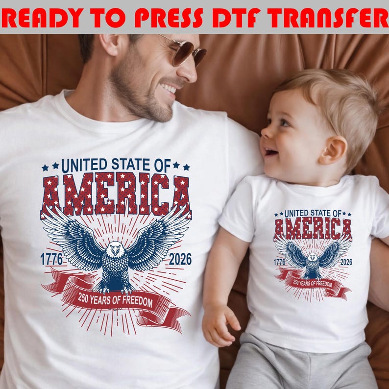 Op de afbeelding: Twee witte t-shirts met een patriottische afbeelding van een Amerikaanse zeearend, met rode en blauwe tekst: UNITED STATE OF AMERICA, 1776, 2026 en 250 YEARS OF FREEDOM. Het ontwerp bevat een rood lint en sterpatronen in de dikke letters.