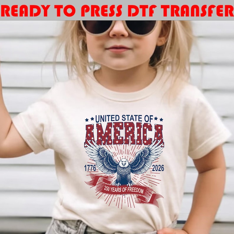 Op de afbeelding: Een cr&egrave;mekleurig t-shirt met een afbeelding van een Amerikaanse zeearend met gespreide vleugels, de tekst UNITED STATE OF AMERICA, 1776, 2026 en 250 YEARS OF FREEDOM. Het ontwerp gebruikt rode, witte en blauwe kleuren met sterpatronen op de letters.