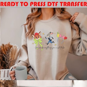 May include: Beige sweatshirt featuring cartoon characters from Zootopia, including Nick Wilde, Judy Hopps, and a Disney Parks headband. The text "You can be anything you wish to be" is below the characters. The words "READY TO PRESS DTF TRANSFER" are at the top.