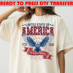 Op de afbeelding: Een off-white t-shirt met een afbeelding van een Amerikaanse zeearend met gespreide vleugels. Boven de adelaar staat in rood en blauw UNITED STATE OF AMERICA. De jaartallen 1776 en 2026 flankeren de adelaar, met een rode banner 250 YEARS OF FREEDOM.