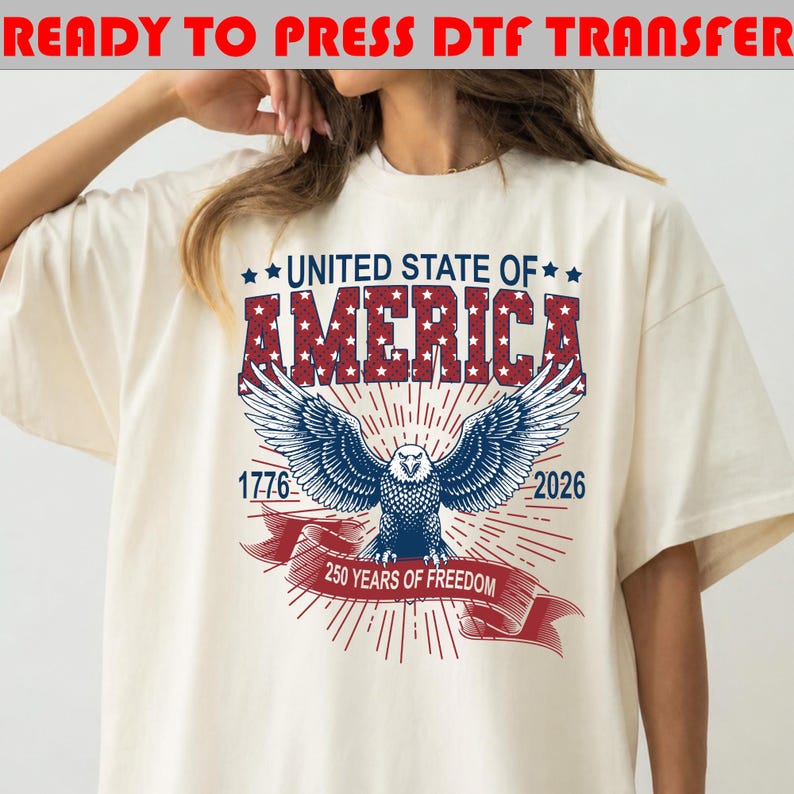 Op de afbeelding: Een off-white t-shirt met een afbeelding van een Amerikaanse zeearend met gespreide vleugels. Boven de adelaar staat in rood en blauw UNITED STATE OF AMERICA. De jaartallen 1776 en 2026 flankeren de adelaar, met een rode banner 250 YEARS OF FREEDOM.