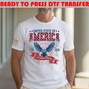 Op de afbeelding: Een wit t-shirt met een afbeelding van een Amerikaanse zeearend met gespreide vleugels, de tekst UNITED STATE OF AMERICA, 1776, 2026 en 250 YEARS OF FREEDOM op een rood lint. De adelaar is blauw en wit met rode sterren op de letters.