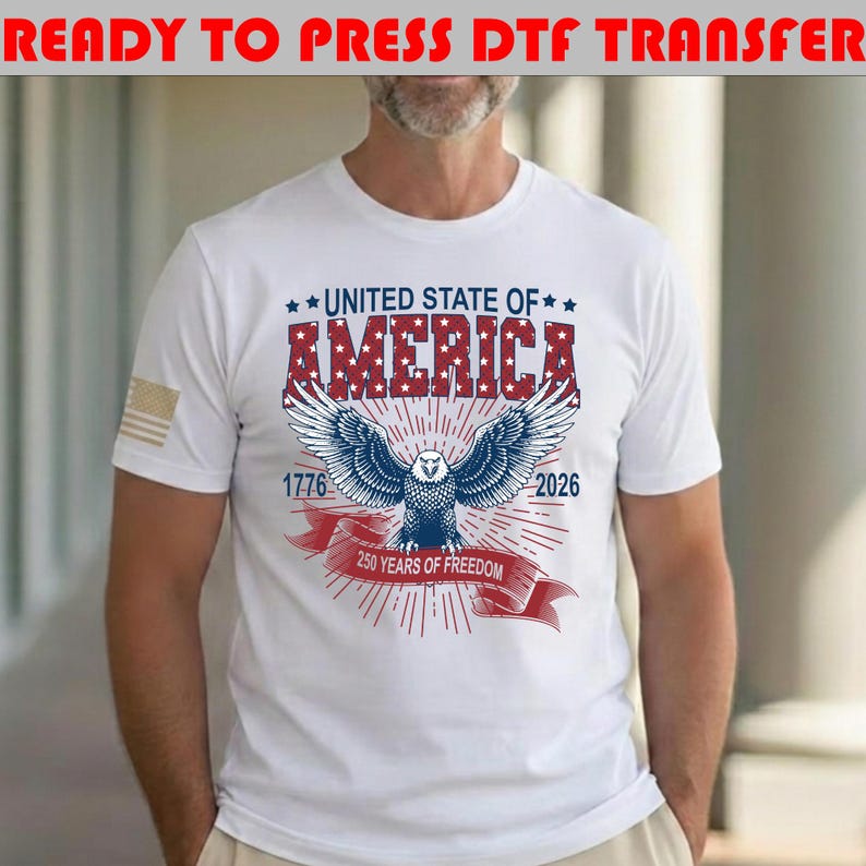 Op de afbeelding: Een wit t-shirt met een afbeelding van een Amerikaanse zeearend met gespreide vleugels, de tekst UNITED STATE OF AMERICA, 1776, 2026 en 250 YEARS OF FREEDOM op een rood lint. De adelaar is blauw en wit met rode sterren op de letters.