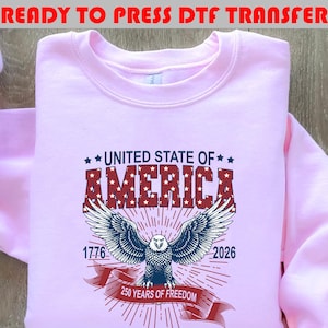 Op de afbeelding: Een lichtroze sweatshirt met een afbeelding met de tekst UNITED STATE OF AMERICA in het rood met witte sterren. Daaronder een blauwe adelaar met gespreide vleugels, de jaartallen 1776 en 2026 en een rode banner met 250 YEARS OF FREEDOM.