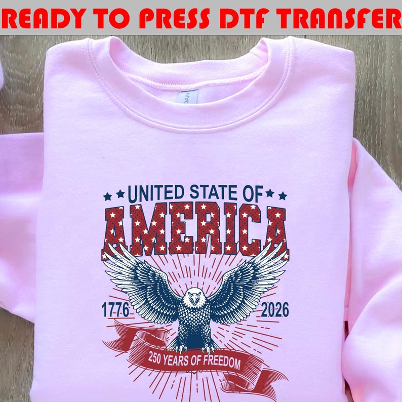 Op de afbeelding: Een lichtroze sweatshirt met een afbeelding met de tekst UNITED STATE OF AMERICA in het rood met witte sterren. Daaronder een blauwe adelaar met gespreide vleugels, de jaartallen 1776 en 2026 en een rode banner met 250 YEARS OF FREEDOM.