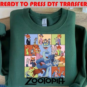 May include: A dark green crewneck sweatshirt featuring a graphic print of various animated characters from Zootopia arranged in a grid. A large blue snake is centered in the foreground. The word Zootopia is printed in black at the bottom of the graphic.