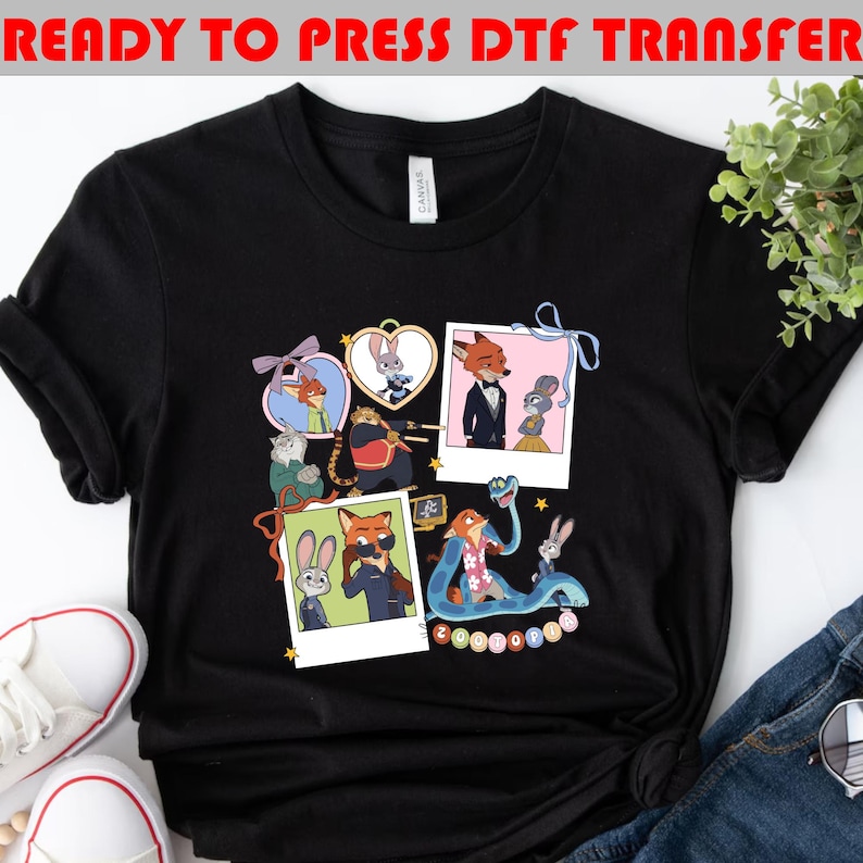 以下が含まれることがあります： ズートピアのキャラクターが描かれた、カラフルな漫画デザインの黒いTシャツ。ニック・ワイルドとジュディ・ホップスのフレーム入り画像が含まれています。「READY TO PRESS DTF TRANSFER」の文字が上部にあります。