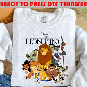 May include: A light gray crewneck sweatshirt featuring a colorful graphic print of characters from The Lion King, including Simba, Mufasa, Scar, Timon, Pumbaa, and Rafiki. The text reads Disney The Lion King in black lettering above the characters.