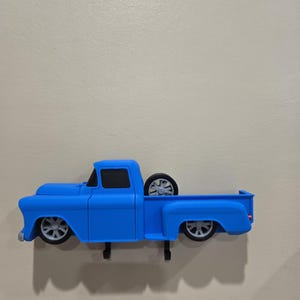 May include: A blue pickup truck-shaped wall hook. The truck has a classic design with a spare tire in the bed and gray wheels. The hook is mounted on a light-colored wall. The truck is approximately 10 inches long.