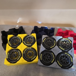 May include: Four sets of toy race car tires in yellow, gray, and red plastic holders. The tires are black with yellow "GOODYEAR EAGLE" lettering. The holders are arranged on a gray surface.