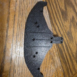 May include: A black, textured, curved metal object with multiple holes and a small rectangular extension. The object has a matte finish and appears to be a component or part of a larger assembly. The shape is reminiscent of a wing or fin.