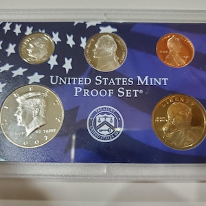 May include: A United States Mint Proof Set in a clear plastic case. The set includes various coins, such as a silver half dollar, a penny, and a dollar coin, against a blue background with white stars and stripes. The text "UNITED STATES MINT PROOF SET" is visible.
