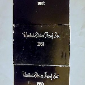 May include: Three stacked black boxes with the text "United States Proof Set" and the years 1980, 1981, and 1982 in a white, elegant font. The boxes are slightly reflective.