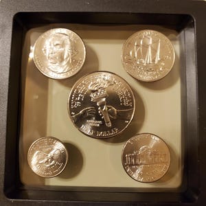 May include: A collection of five US coins displayed in a black frame. The coins feature various designs, including portraits, ships, and the torch of liberty. The coins are in excellent condition and are set against a neutral background.