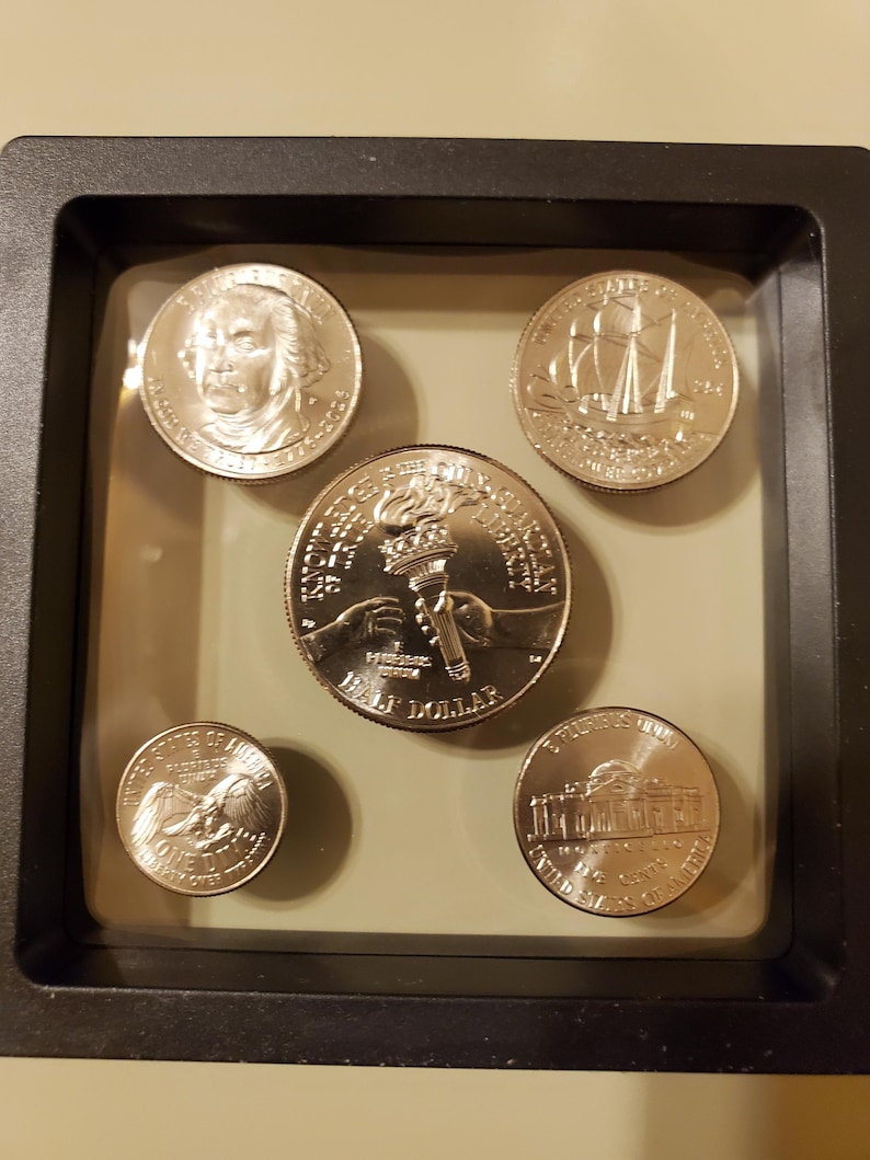 May include: A collection of five US coins displayed in a black frame. The coins feature various designs, including portraits, ships, and the torch of liberty. The coins are in excellent condition and are set against a neutral background.