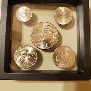May include: A collection of five silver coins displayed in a black shadow box. The coins feature portraits and historical dates. The largest coin depicts the Statue of Liberty. The coins are encased in a clear, protective display.