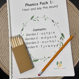 May include: A white spiral-bound Phonics Pack 1 workbook with the text Hear and say the sound and a list of contents labelled Garden 1 through 4. A small brown box of coloured pencils and a single yellow pencil with an eraser are placed on top.