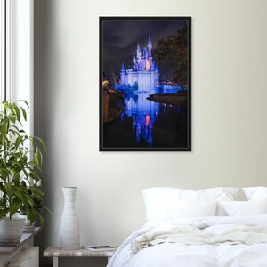 May include: A framed print of a fairytale castle at night, illuminated with blue and purple lights. The castle is reflected in the water below, creating a symmetrical image. The artwork is displayed on a white wall.