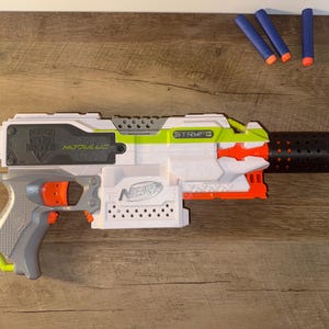 May include: A white, grey, and orange Nerf Modulus Stryfe toy gun with a black barrel extension. The toy gun has the word "Nerf" printed on the side and comes with three blue and orange darts.