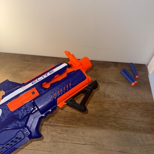 May include: A blue and orange toy blaster with the word "ELITE" printed on the side. The blaster has a black handle and several blue foam darts are next to it. The blaster is on a wooden surface.