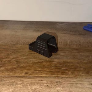 Op de afbeelding: Een zwarte, 3D-geprinte vizierbevestiging voor een speelgoedpistool, met horizontale lijnen en rechthoekige details. Het vizier ligt op een houten oppervlak, met blauwe en oranje speelgoedpijlen op de achtergrond.