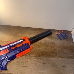 May include: A blue and orange toy blaster with a black barrel extension and three blue and orange darts. The blaster has the word "ELITE" printed on the side. The toy is on a wooden surface.