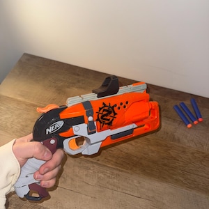 Op de afbeelding: Een oranje en grijze Nerf gun met een zwart vizier en het woord "NERF" gedrukt op de zijkant. Het pistool heeft een zwarte en rode handgreep en een grafisch ontwerp aan de zijkant. Drie blauwe darts liggen naast het pistool.