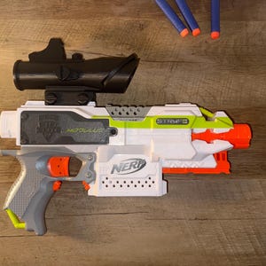 Op de afbeelding: Een witte, grijze en oranje Nerf Stryfe Modulus speelgoedpistool met een zwarte richtkijker. Het speelgoedpistool heeft een grijze handgreep en oranje accenten. Extra blauwe en oranje darts zijn zichtbaar.