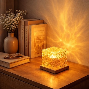 Puede incluir: Una lámpara cúbica decorativa con una pantalla de vidrio ámbar texturizado se encuentra sobre una base de madera. La lámpara está sobre una superficie de madera, junto a libros y un jarrón con flores blancas. La luz proyecta un brillo cálido y estampado en la pared.
