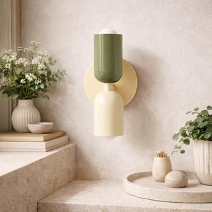 May include: A modern wall sconce with a two-tone design. The top section is olive green, the middle is a pale yellow circle, and the bottom is a matching pale yellow. The sconce is mounted on a textured wall, with a small light bulb visible.