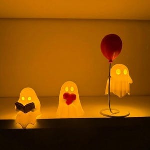 May include: Three illuminated ghost-shaped lamps, perfect for Halloween. One ghost reads a book, another has a red heart, and the third holds a red balloon. The lamps emit a warm, yellow glow, creating a cosy and festive ambiance.