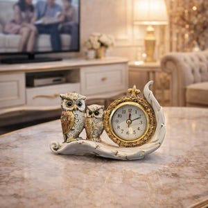 May include: A decorative owl clock featuring two owl figurines and a clock face. The owls are in shades of brown, white, and gold. The clock has a gold frame and a white base. The clock is on a marble table.