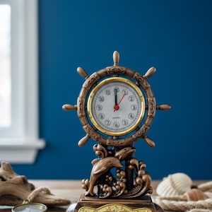 Könnte beinhalten: Eine Uhr im nautischen Stil mit einem Schiffsrad-Design in Bronzefarbe. Das Zifferblatt ist weiß mit schwarzen Ziffern und roten Zeigern. Am Sockel sind zwei Delfine abgebildet. Die Uhr ist etwa 15 cm hoch.